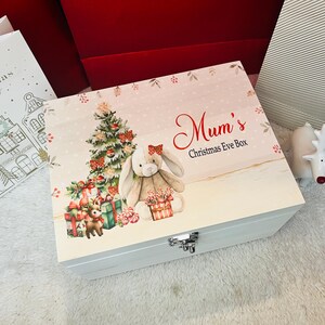 May include: A white wooden box with a hinged lid, decorated with a Christmas Eve theme. The lid features a Christmas tree, a plush bunny, and the text "Mum's Christmas Eve Box". The box is adorned with gifts and candy canes.