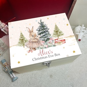 Puede incluir: Una caja rectangular de madera con tapa abatible que presenta una ilustración en acuarela de un conejo con tutú junto a árboles de Navidad y regalos. El texto en la tapa dice Alice's Christmas Eve Box. La caja tiene un cierre de metal plateado.