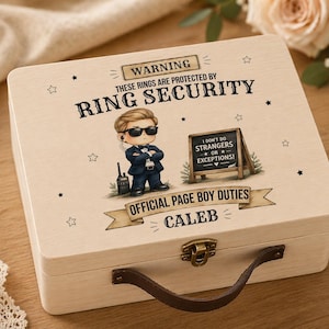 May include: A wooden ring box with a brown leather handle and metal clasp. The box features a cartoon illustration of a security guard with the text "WARNING THESE RINGS ARE PROTECTED BY RING SECURITY" and "OFFICIAL PAGE BOY DUTIES CALEB".