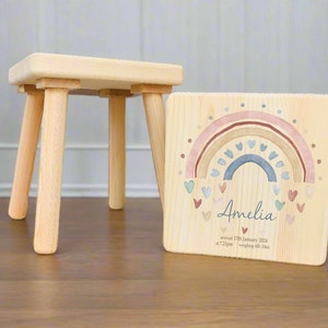 Puede incluir: Un taburete de madera con un diseño de arcoíris y corazón en el lateral. El diseño incluye el nombre Amelia, la fecha 12 de enero de 2024 y la hora 7:21 pm. El texto también dice "pesando 6lb 10oz".