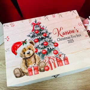 Puede incluir: Una caja de madera rectangular con una ilustración impresa de un oso de peluche marrón con un gorro de Papá Noel rojo, un árbol de Navidad decorado con bolas rojas y regalos. El texto dice Karson's Christmas Eve box 2025.