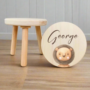 May include: A wooden stool with three legs and a round top. The top is painted with a lion face and the name "George".