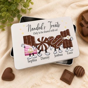 以下が含まれることがあります： Nandad's TreatsおよびOnly to be shared with usというテキストが書かれた長方形の白い金属製の缶。蓋には、Sophia、Darcey、Joshua、Lucaという名前が下に印刷された、擬人化された4つのチョコレートキャラクターが描かれています。