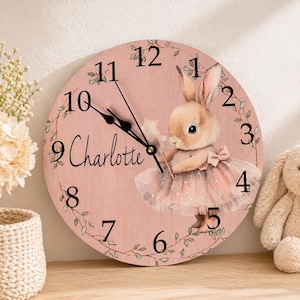 Könnte beinhalten: Eine runde, rosa Holz-Uhr mit schwarzen Ziffern und Zeigern. Das Zifferblatt zeigt eine Aquarell-Illustration eines Häschens in einem Tutu, das einen Zauberstab hält. Der Name "Charlotte" ist kursiv geschrieben. Die Uhr ist von einem grünen Rankenmuster umgeben.