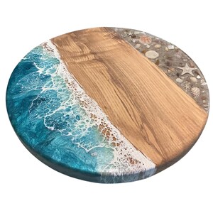May include: Round wooden serving board with a beach-themed design. The board features a section of light brown wood, a section of blue resin simulating ocean waves, and a section with seashells and sand.