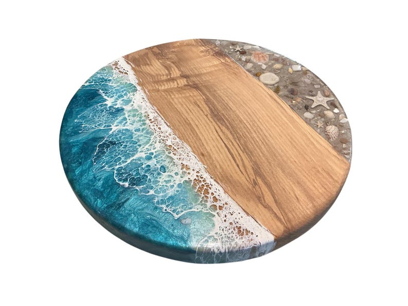 May include: Round wooden serving board with a beach-themed design. The board features a section of light brown wood, a section of blue resin simulating ocean waves, and a section with seashells and sand.