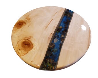 May include: Oval wooden serving board with a river of blue resin and embedded stones. The light-colored wood has natural knots and grain patterns. The resin river runs down the center of the board, creating a unique decorative piece.