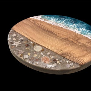 May include: Oval wooden serving board with a beach-themed design. The board features a section of light-colored wood, a sandy area with seashells and starfish, and a blue and white resin wave design. The board is set against a black background.