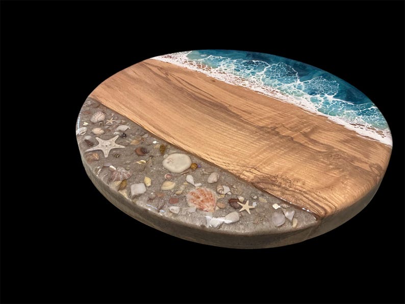 May include: Oval wooden serving board with a beach-themed design. The board features a section of light-colored wood, a sandy area with seashells and starfish, and a blue and white resin wave design. The board is set against a black background.
