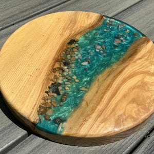 May include: A round wooden serving board with a river of teal resin and embedded stones. The wood has a light brown color with visible grain. The resin river runs through the center of the board, creating a unique, natural design.