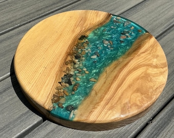 May include: A round wooden serving board with a river of teal resin and embedded stones. The wood has a light brown color with visible grain. The resin river runs through the center of the board, creating a unique, natural design.