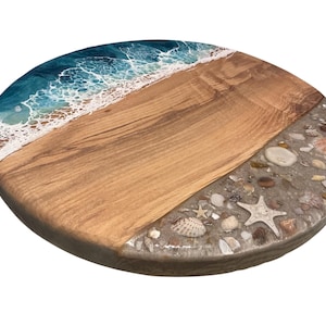 May include: A round wooden serving board with a beach-themed design. The board features a section of light brown wood, a section with seashells and sand, and a blue and white resin wave design. The board is perfect for serving appetizers.