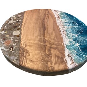 May include: Oval wooden serving board with a beach-themed design. One section features embedded seashells and sand, another has natural wood grain, and the third depicts ocean waves in shades of blue and white.