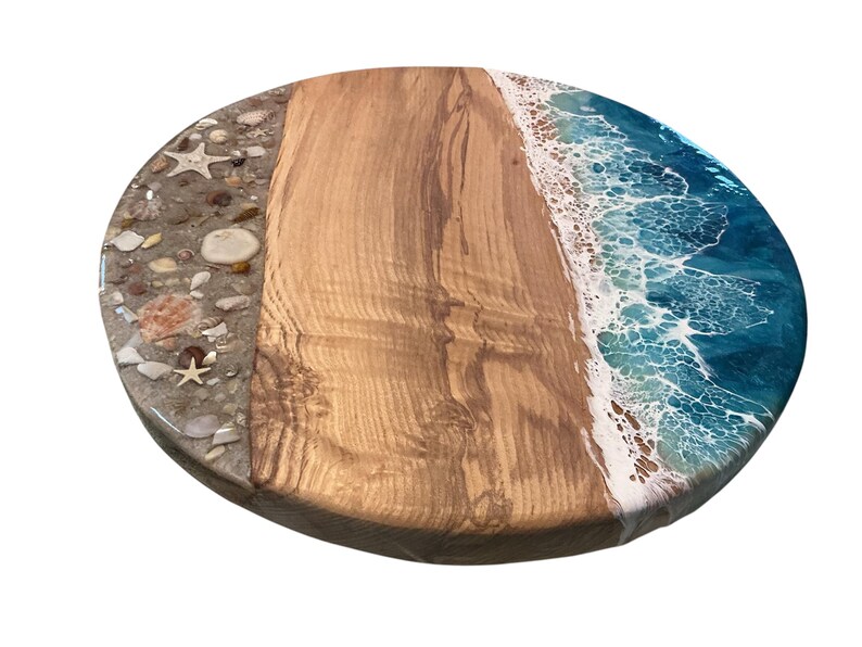 May include: Oval wooden serving board with a beach-themed design. One section features embedded seashells and sand, another has natural wood grain, and the third depicts ocean waves in shades of blue and white.