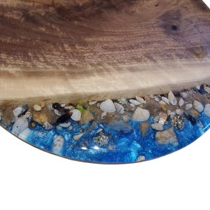 Könnte beinhalten: Rundes Holzservierbrett mit Strandmotiv. Das Brett hat eine natürliche Holzmaserung, einen sandigen Strandabschnitt mit Muscheln und Kieselsteinen und einen leuchtend blauen Harz-Ozean.
