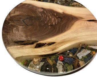 May include: A round wooden serving board with a natural wood grain pattern. The board features a clear resin river with stones, moss, and a red mushroom. The wood is light brown and dark brown.