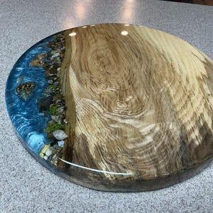 May include: A round wooden serving board with a river design. The board features a section of natural wood grain and a section of blue resin with embedded stones, creating a river effect. The board is approximately 16 inches in diameter.