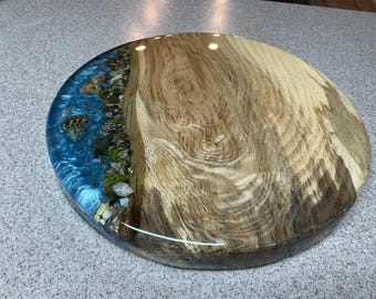 May include: A round wooden serving board with a river design. The board features a section of natural wood grain and a section of blue resin with embedded stones, creating a river effect. The board is approximately 16 inches in diameter.