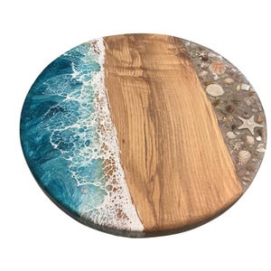 May include: Round wooden serving board with a beach-themed design. The board features a section of light brown wood, a turquoise and white resin wave design, and a border with seashells and sand.