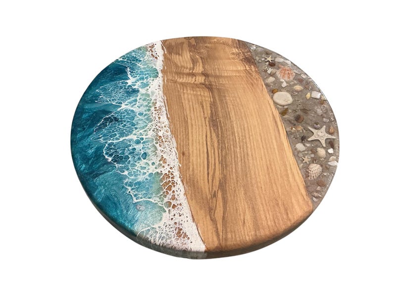 May include: Round wooden serving board with a beach-themed design. The board features a section of light brown wood, a turquoise and white resin wave design, and a border with seashells and sand.