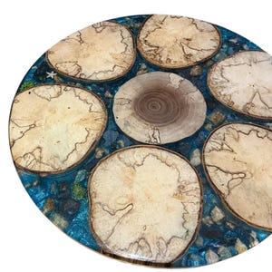 May include: A circular wooden table with a distinctive design. It has a blue resin base with embedded stones and wood slices. The wood slices are light brown with detailed patterns. The table's diameter is approximately 61 cm.