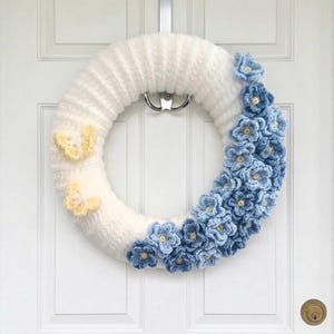 May include: A circular wreath wrapped in white yarn, featuring a cluster of crocheted blue flowers along the right side and two small, pale yellow crocheted butterflies attached to the left side, hanging from a silver metal door hook.