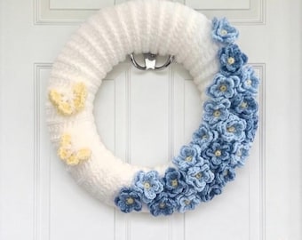 Könnte beinhalten: Ein kreisförmiger Kranz, der mit weißem Garn umwickelt ist, mit einer Ansammlung gehäkelter blauer Blumen auf der rechten Seite und zwei kleinen, hellgelben gehäkelten Schmetterlingen auf der linken Seite, hängend an einem silbernen Metallhaken.