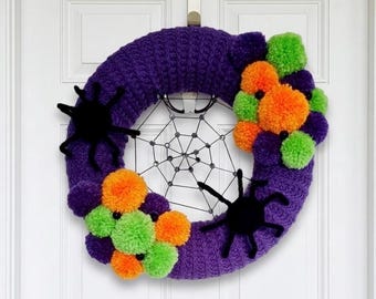 Könnte beinhalten: Ein kreisförmiger Kranz aus violettem Garn mit einem schwarzen Spinnennetz-Design in der Mitte. Der Kranz ist mit Büscheln aus leuchtend orangefarbenen und lindgrünen Pompons sowie zwei schwarzen Spinnenfiguren an den Seiten verziert.