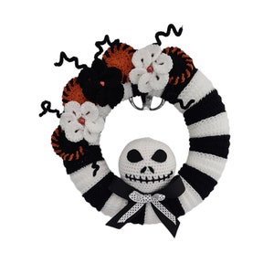 Op de afbeelding: Een zwart-wit gestreepte gehaakte krans met oranje en witte bloemen en een wit gehaakt Jack Skellington hoofd in het midden.