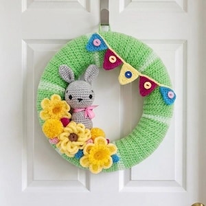 May include: A crochet wreath featuring a light green base with a grey bunny figure, yellow flowers, and a colourful bunting banner with small buttons. The bunny has a pink ribbon around its neck, and the flowers are arranged in a cluster at the bottom.
