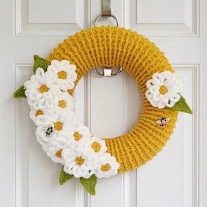 Puede incluir: Una corona circular con una base de ganchillo de color amarillo mostaza texturizado, adornada con varias flores de margarita de ganchillo blancas con centros amarillos, hojas de ganchillo verdes y dos pequeñas abejas decorativas.