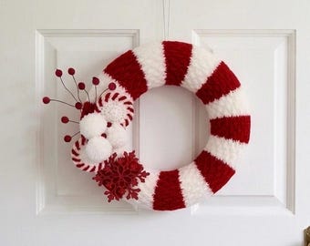 May include: A circular wreath wrapped in alternating red and white yarn stripes. The left side is decorated with three white yarn pom-poms, two small red and white striped circular accents, a red glittery snowflake, and thin wire stems with red berries.
