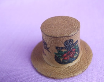 Puede incluir: Un pequeño sombrero de cuero marrón con un diseño de ancla negra y blanca y una flor roja.