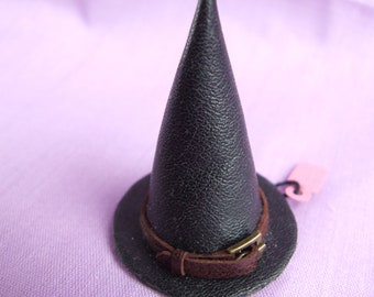 Puede incluir: Un sombrero de bruja negro con una banda de cuero marrón y una hebilla. El sombrero está sobre un fondo morado.
