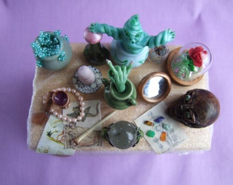 Puede incluir: Una mesa en miniatura con una criatura verde y azul, una cúpula de vidrio con una rosa roja, una bola de cristal, una pequeña mano verde, un collar de perlas, un pequeño cuenco con cuentas azules y una calavera.