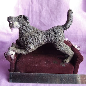 May include: A gray, furry dog figurine is posed on a burgundy velvet couch. The dog is standing on its hind legs with its front paws resting on the edge of the couch. The figurine is approximately 15 cm long.