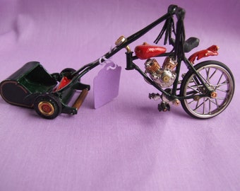 Puede incluir: Motocicleta en miniatura con sidecar, con un marco negro, depósito de combustible rojo y componentes del motor plateados. El sidecar es verde oscuro con detalles dorados. Una etiqueta púrpura en blanco cuelga del manillar. Tiene una rueda trasera.