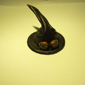 May include: A black leather witch hat with a pointed crown and a brim. The hat has two round, orange goggles attached to the brim.
