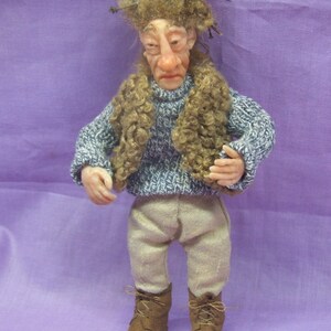 May include: A handcrafted doll depicting a whimsical character. The doll wears a blue and white sweater, tan pants, and brown boots. It has a detailed face with a prominent nose and curly brown hair. The doll is approximately 6 inches tall.
