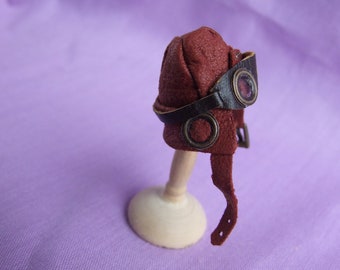 Puede incluir: Un casco de aviador de cuero burdeos en miniatura con gafas y correas de cuero marrón oscuro. El casco se exhibe sobre un pequeño soporte de madera sobre un fondo púrpura. Las gafas tienen lentes circulares.