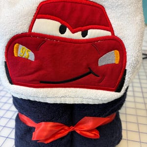 May include: A white hooded towel featuring an embroidered red race car with white eyes, a black mouth, and the number 95 in yellow on the side. The towel is rolled and secured with a red satin ribbon tied in a bow around the dark blue base.