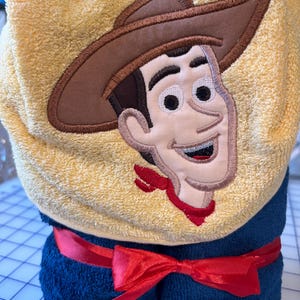 May include: A yellow hooded towel with an embroidered image of a cartoon cowboy wearing a brown hat and a red bandana. The towel is rolled up and tied with a red ribbon.