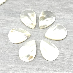 May include: A collection of teardrop-shaped mother of pearl cabochons with a smooth, iridescent finish. The pieces are white and off-white with natural pearlescent luster, arranged in a scattered pile with six pieces placed in a flower pattern.