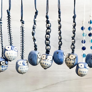 May include: A collection of ceramic owl pendants in various styles. Each owl is blue and white, with detailed facial features. The pendants hang from cords with decorative knots and beads, creating a charming and whimsical aesthetic.