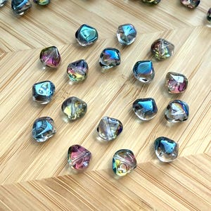 May include: Iridescent glass beads with a faceted, irregular shape. The beads reflect a spectrum of colors, including blues, greens, pinks, and purples. These beads are scattered on a light brown, wooden surface. Ideal for jewelry making and crafts.