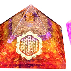 May include: A clear crystal pyramid with a gold flower of life design and a copper coil inside. The pyramid is filled with orange, red, and purple crystals. A purple crystal pendulum hangs on a gold chain.
