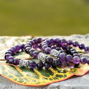 May include: Three amethyst gemstone bracelets with silver Buddha head beads. The bracelets are stacked on a green and yellow leaf.