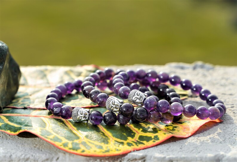 May include: Three amethyst gemstone bracelets with silver Buddha head beads. The bracelets are stacked on a green and yellow leaf.