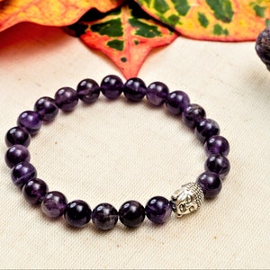 May include: A purple amethyst gemstone bracelet with a silver Buddha head charm. The bracelet is made of round beads and is strung on an elastic cord.