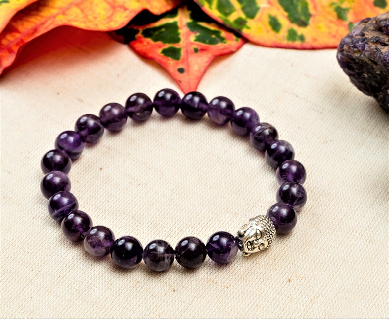 May include: A purple amethyst gemstone bracelet with a silver Buddha head charm. The bracelet is made of round beads and is strung on an elastic cord.